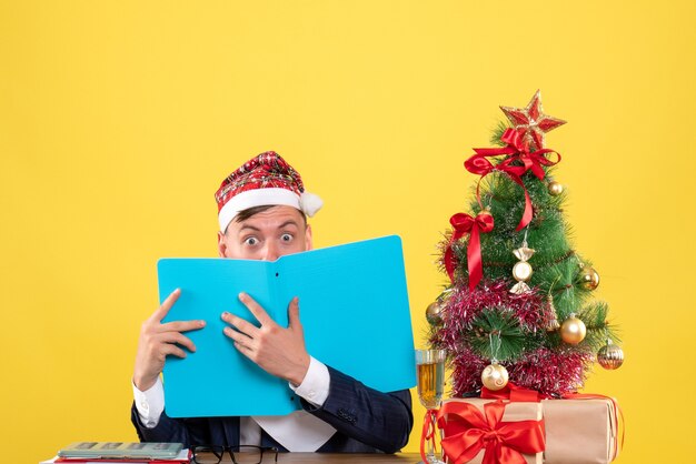 Vue de face de l'homme d'affaires aux yeux écarquillés assis à la table près de l'arbre de Noël et présente sur jaune