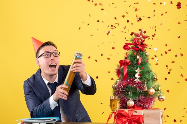 Vue de face de l'homme d'affaires avec capuchon à l'aide de partie popper debout derrière la table près de l'arbre de Noël et présente sur jaune