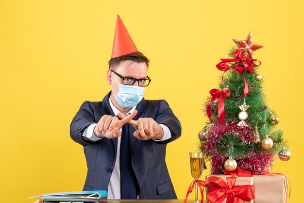 Vue de face homme d'affaires croisant les doigts assis à la table près de l'arbre de Noël et présente sur fond jaune