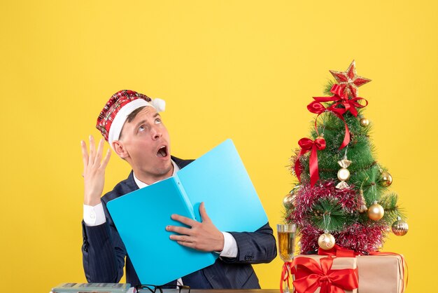 Vue de face de l'homme d'affaires détenant des documetns assis à la table près de l'arbre de Noël et présente sur jaune