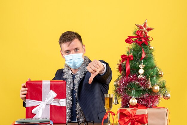 Vue de face de l'homme d'affaires faisant le pouce vers le bas signe assis à la table près de l'arbre de Noël et présente sur jaune