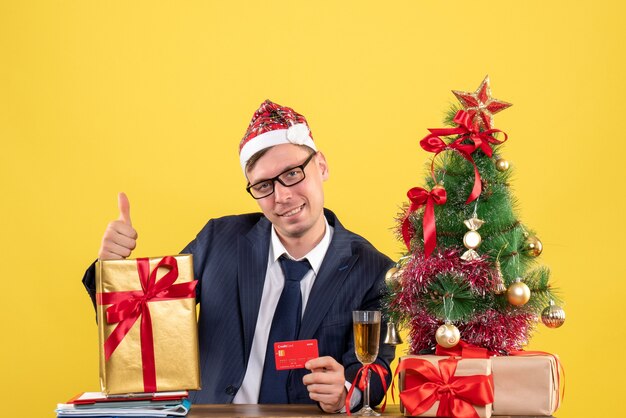 Vue de face de l'homme d'affaires faisant le pouce vers le haut signe assis à la table près de l'arbre de Noël et présente sur jaune