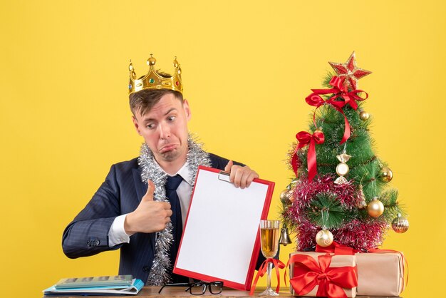 Vue de face de l'homme d'affaires faisant le pouce vers le haut signe assis à la table près de l'arbre de Noël et présente sur jaune