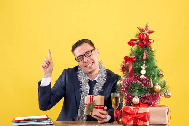 Vue de face de l'homme d'affaires heureux doigt pointé vers le haut assis à la table près de l'arbre de Noël et présente sur jaune