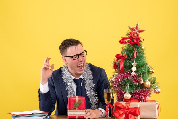 Vue de face de l'homme d'affaires heureux faisant signe de bonne chance assis à la table près de l'arbre de Noël et présente sur jaune