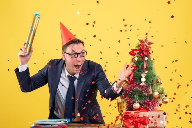 Vue de face de l'homme d'affaires holding party popper debout derrière la table près de l'arbre de Noël et présente sur jaune