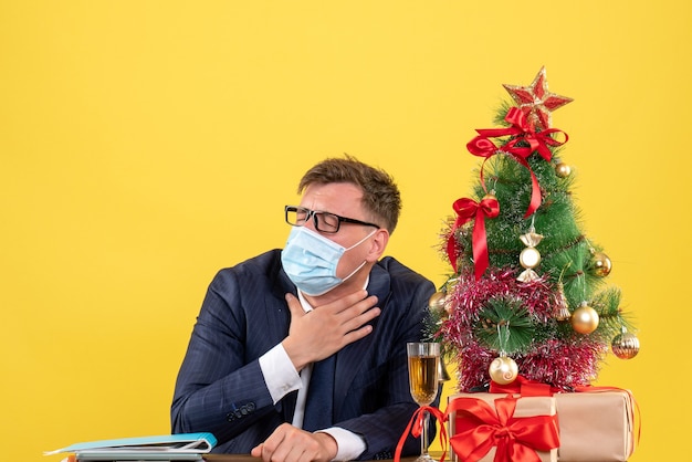 Vue de face de l'homme d'affaires avec masque fermant les yeux assis à la table près de l'arbre de Noël et présente sur jaune