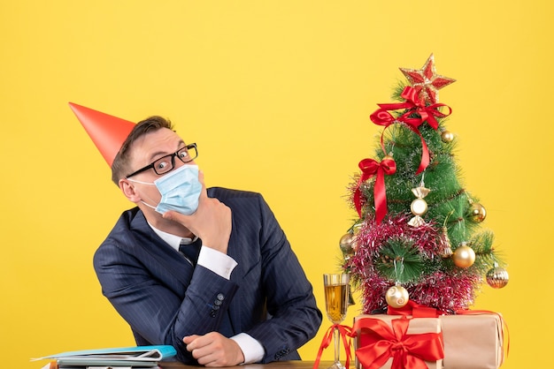 Vue De Face De L'homme D'affaires Mettant La Main Sur Son Menton Assis à La Table Près De L'arbre De Noël Et Présente Sur Jaune