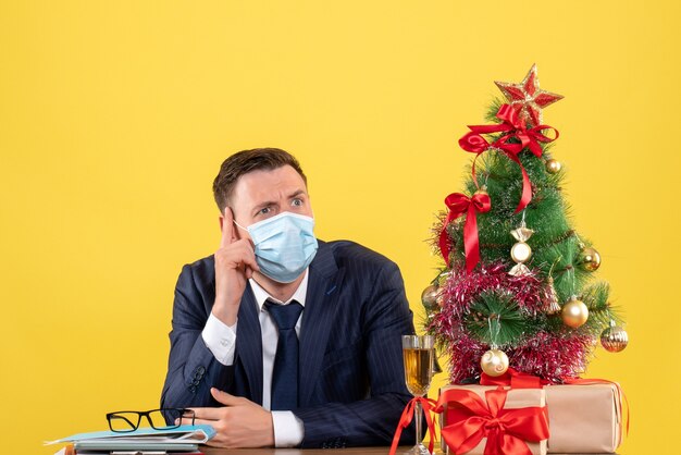 Vue de face de l'homme d'affaires mettant son doigt sur son temple assis à la table près de l'arbre de Noël et présente sur jaune