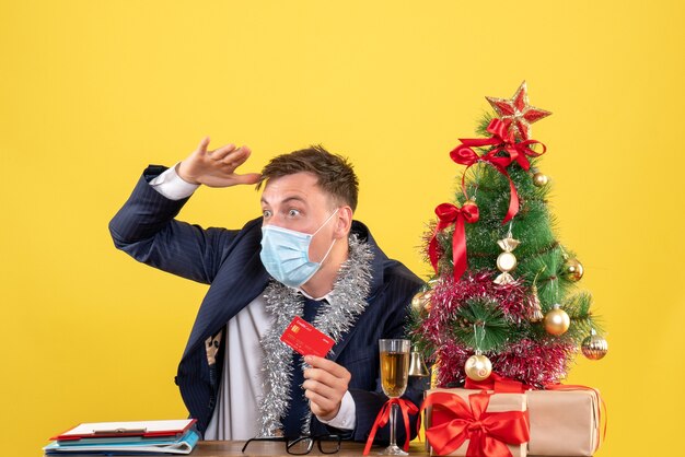 Vue de face de l'homme d'affaires observant quelque chose assis à la table près de l'arbre de Noël et présente sur jaune