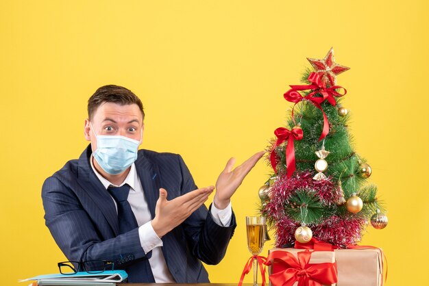 Vue de face de l'homme d'affaires pointant sur l'arbre de Noël assis à la table près de l'arbre de Noël et présente sur jaune