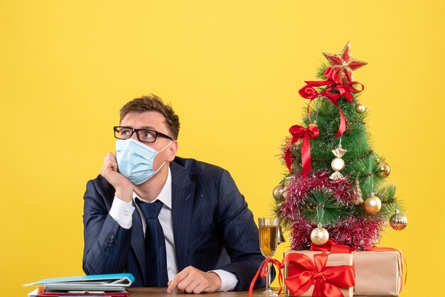 Vue de face de l'homme d'affaires à la recherche assis à la table près de l'arbre de Noël et présente sur jaune