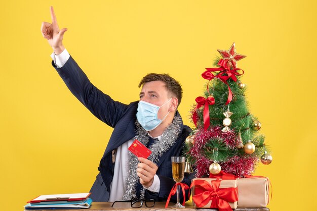 Vue de face de l'homme d'affaires regardant quelque chose assis à la table près de l'arbre de Noël et présente sur jaune