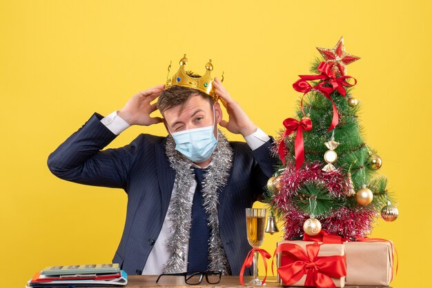 Vue de face de l'homme d'affaires tenant la couronne assis à la table près de l'arbre de Noël et présente sur jaune