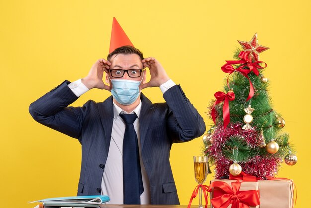 Vue de face de l'homme d'affaires tenant sa tête assise à la table près de l'arbre de Noël et présente sur jaune