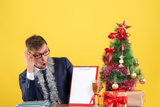 Vue de face de l'homme d'affaires vérifiant le papier assis à la table près de l'arbre de Noël et présente sur fond jaune