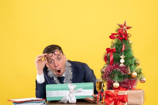 Vue de face de l'homme aux yeux écarquillés qui décolle des lunettes assis à la table près de l'arbre de Noël et présente sur jaune