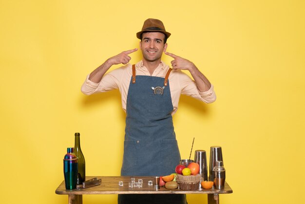 Vue de face homme barman debout devant le bureau avec des boissons et des fruits souriant sur un mur jaune boisson night club bar à jus homme