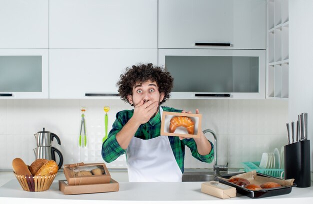 Vue de face d'un homme choqué tenant une pâtisserie fraîchement préparée dans une petite boîte dans la cuisine blanche