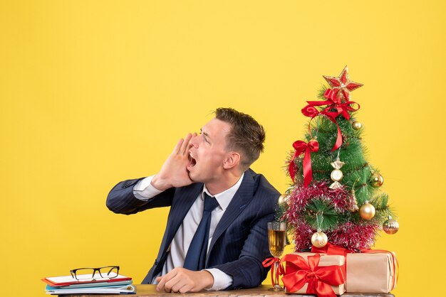 Photo gratuite vue de face de l'homme en colère appelant quelqu'un assis à la table près de l'arbre de noël et des cadeaux sur le mur jaune