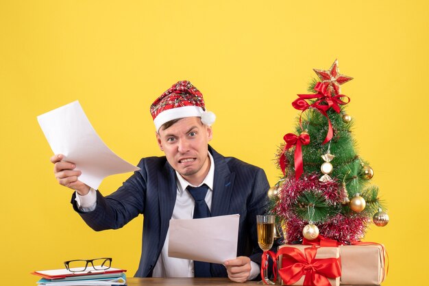 Vue de face de l'homme en colère avec Bonnet de Noel assis à la table près de l'arbre de Noël et présente sur l'espace de copie de mur jaune