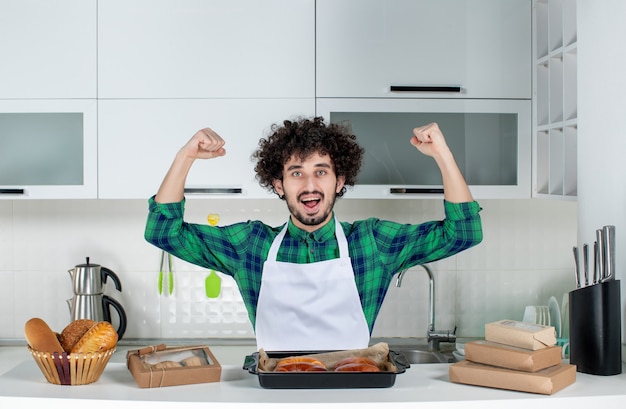 Vue De Face D'un Homme Confiant Debout Derrière Une Table Avec Une Pâtisserie Fraîchement Cuite Dessus Et Montrant Ses Muscles Dans La Cuisine Blanche