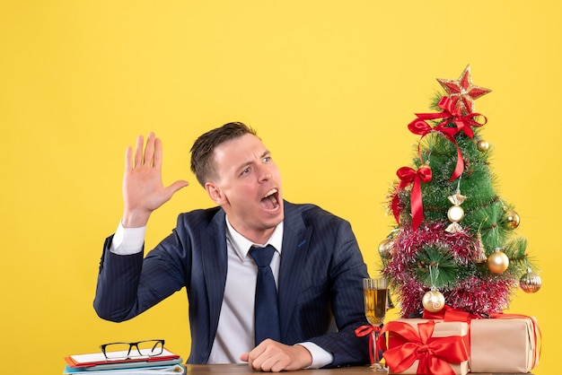 Vue de face de l'homme en costume saluant quelqu'un assis à la table près de l'arbre de Noël et des cadeaux sur jaune