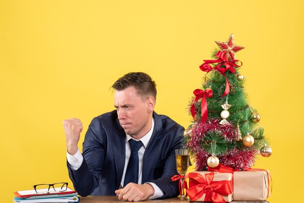 Vue de face de l'homme en costume se regardant assis à la table près de l'arbre de Noël et des cadeaux sur jaune