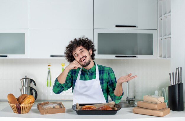 Vue de face d'un homme curieux debout derrière une table avec une pâtisserie fraîchement préparée et faisant un geste de l'appel dans la cuisine blanche