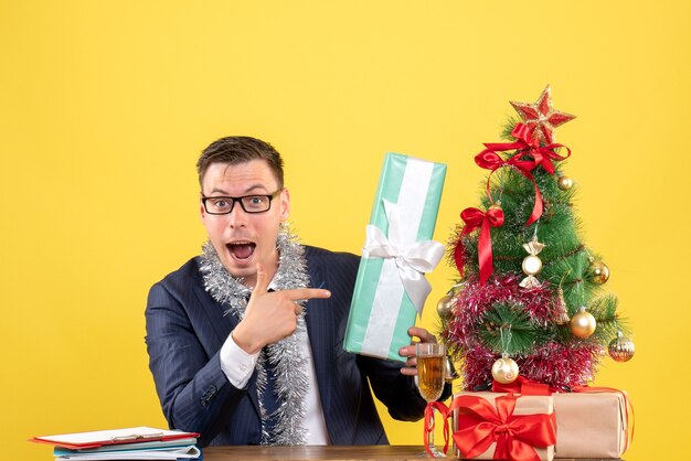 Vue de face de l'homme demandé, pointant sur cadeau assis à la table près de l'arbre de Noël et présente sur jaune