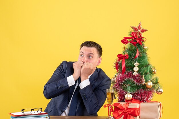 Vue de face de l'homme demandé à quelque chose assis à la table près de l'arbre de Noël et cadeaux sur jaune