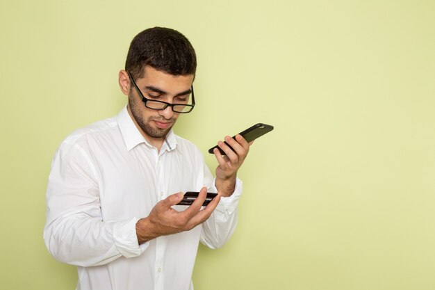 Vue de face de l'homme employé de bureau en chemise blanche tenant et à l'aide de son téléphone sur le mur vert clair