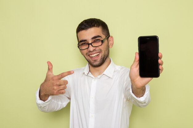 Vue de face de l'homme employé de bureau en chemise blanche tenant son téléphone souriant sur mur vert