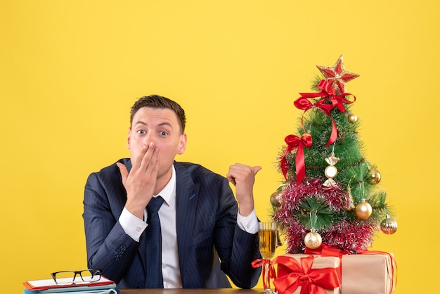 Vue de face homme étonné de pointage du doigt arbre de Noël assis à la table près de l'arbre de Noël et des cadeaux sur fond jaune