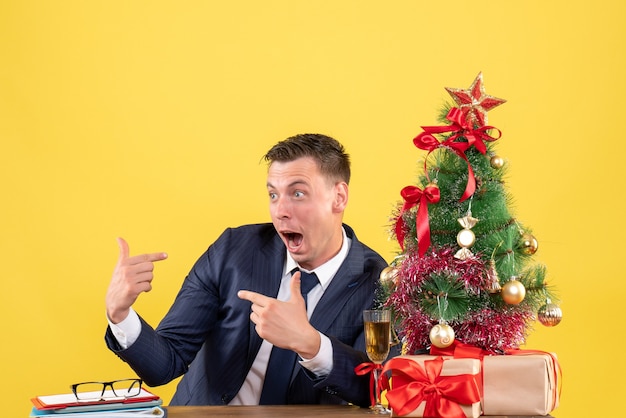 Vue de face de l'homme étonné se montrant assis à la table près de l'arbre de Noël et des cadeaux sur le mur jaune