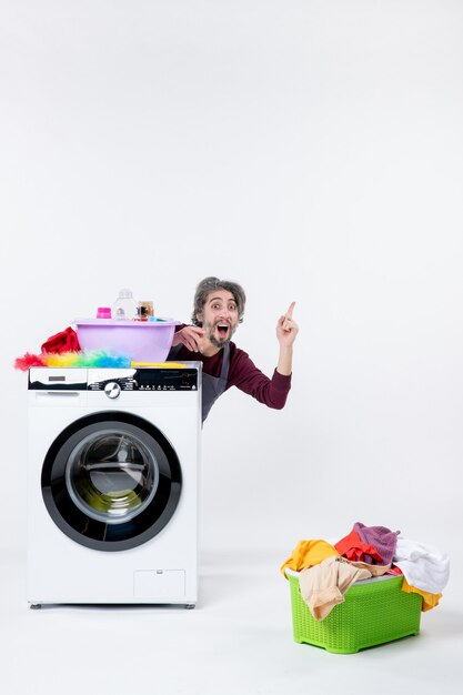 Vue de face homme étonné en tablier assis derrière le panier à linge lave-linge sur fond isolé blanc