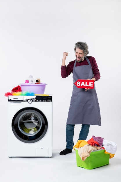 Photo gratuite vue de face homme exalté en tablier brandissant une pancarte de vente debout près de la laveuse sur fond blanc