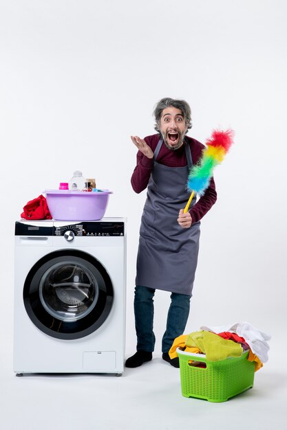 Vue de face homme de femme de ménage étonné tenant duster debout près du panier à linge de la machine à laver sur fond blanc