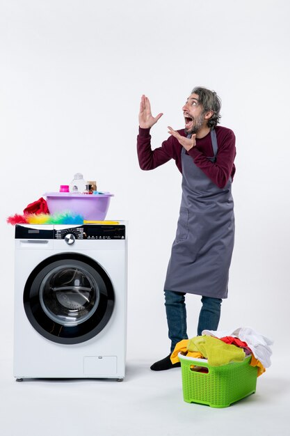 Vue de face homme de femme de ménage excité regardant debout près du panier à linge de la laveuse sur fond blanc