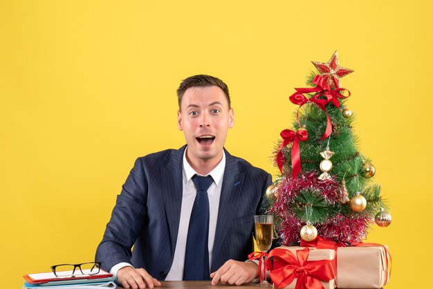 Vue de face de l'homme heureux assis à la table près de l'arbre de Noël et des cadeaux sur jaune