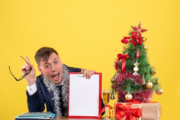 Vue de face de l'homme heureux assis à la table près de l'arbre de Noël et présente sur jaune