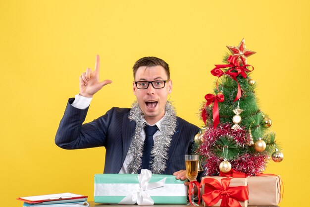 Vue de face de l'homme heureux doigt pointé vers le haut assis à la table près de l'arbre de Noël et présente sur jaune