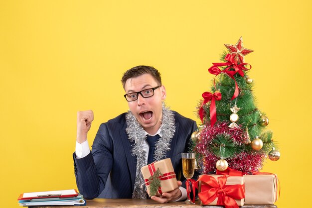 Vue de face de l'homme heureux avec des lunettes assis à la table près de l'arbre de Noël et présente sur jaune