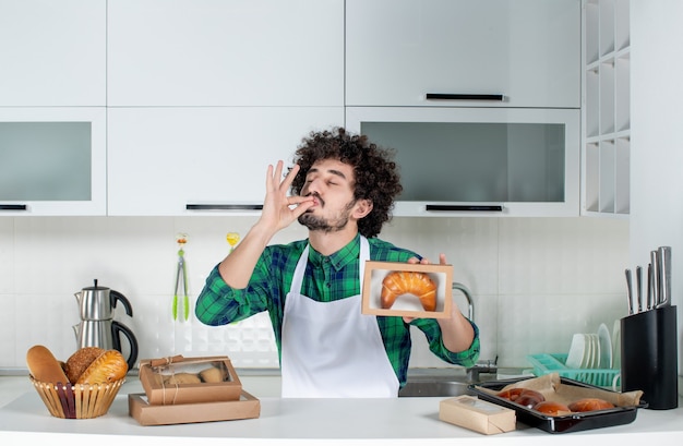 Vue de face d'un homme heureux tenant une pâtisserie fraîchement préparée dans une petite boîte et faisant un geste parfait dans la cuisine blanche
