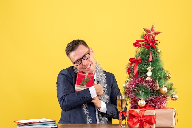 Vue de face de l'homme heureux tenant son cadeau étroitement assis à la table près de l'arbre de Noël et présente sur jaune