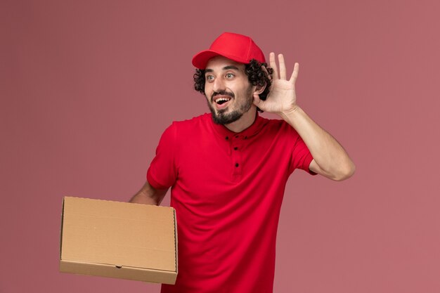 Vue de face homme livreur de courrier en chemise rouge et cape tenant la boîte de nourriture de livraison sur le mur rose service de livraison emploi employé de l'entreprise