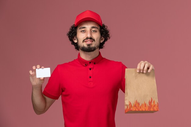 Vue de face homme livreur de courrier en chemise rouge et cape tenant le paquet de nourriture et la carte sur le mur rose employé de livraison de services