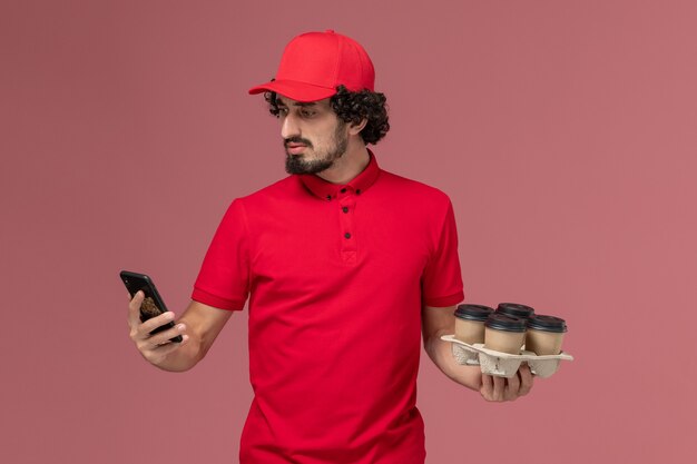 Vue de face homme livreur de courrier en chemise rouge et cape tenant des tasses de café de livraison marron avec téléphone sur le mur rose clair employé de livraison de service d'emploi