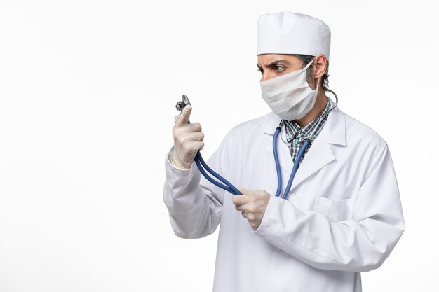 Vue de face de l'homme médecin en costume médical blanc et masque en raison du coronavirus tenant un stéthoscope sur une surface blanche claire