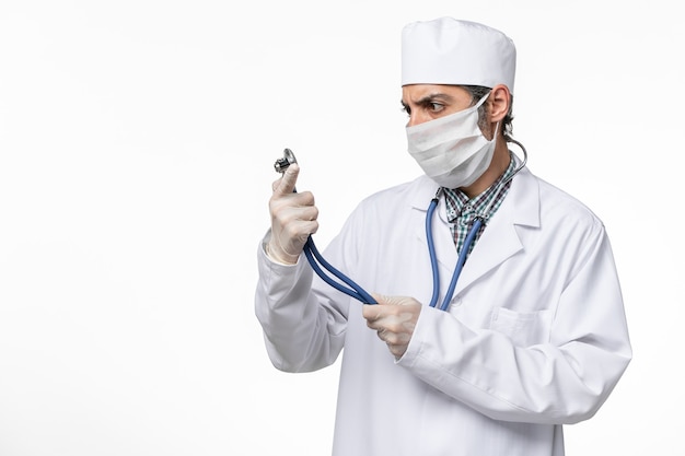 Vue de face de l'homme médecin en costume médical blanc et masque en raison du coronavirus tenant un stéthoscope sur une surface blanche claire